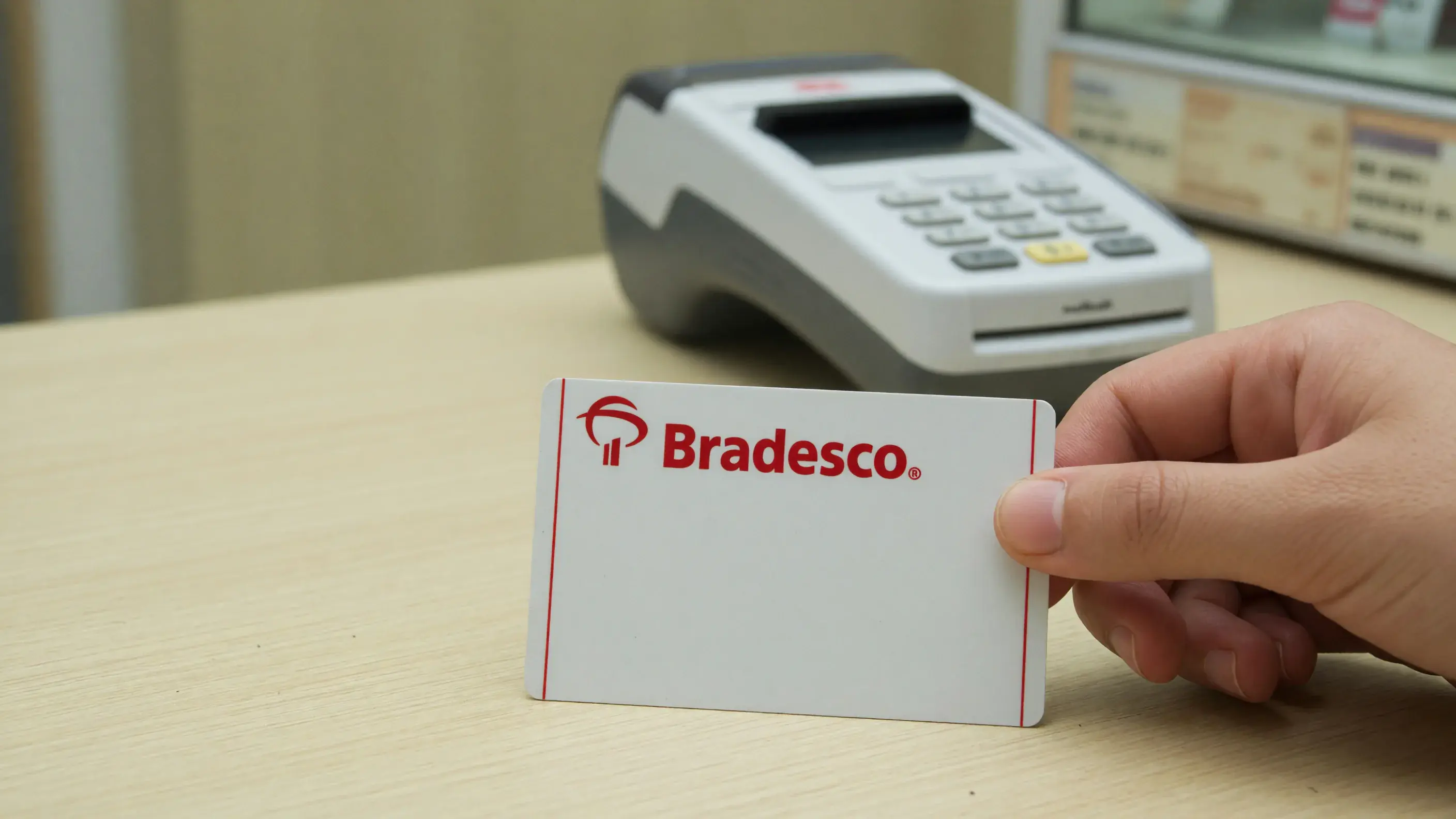 Cartão de crédito Bradesco na mão de um cliente sobre o balcão de uma loja, com a marca do banco nítida na face do cartão, sem nenhum número impresso, e a maquininha desfocada ao lado