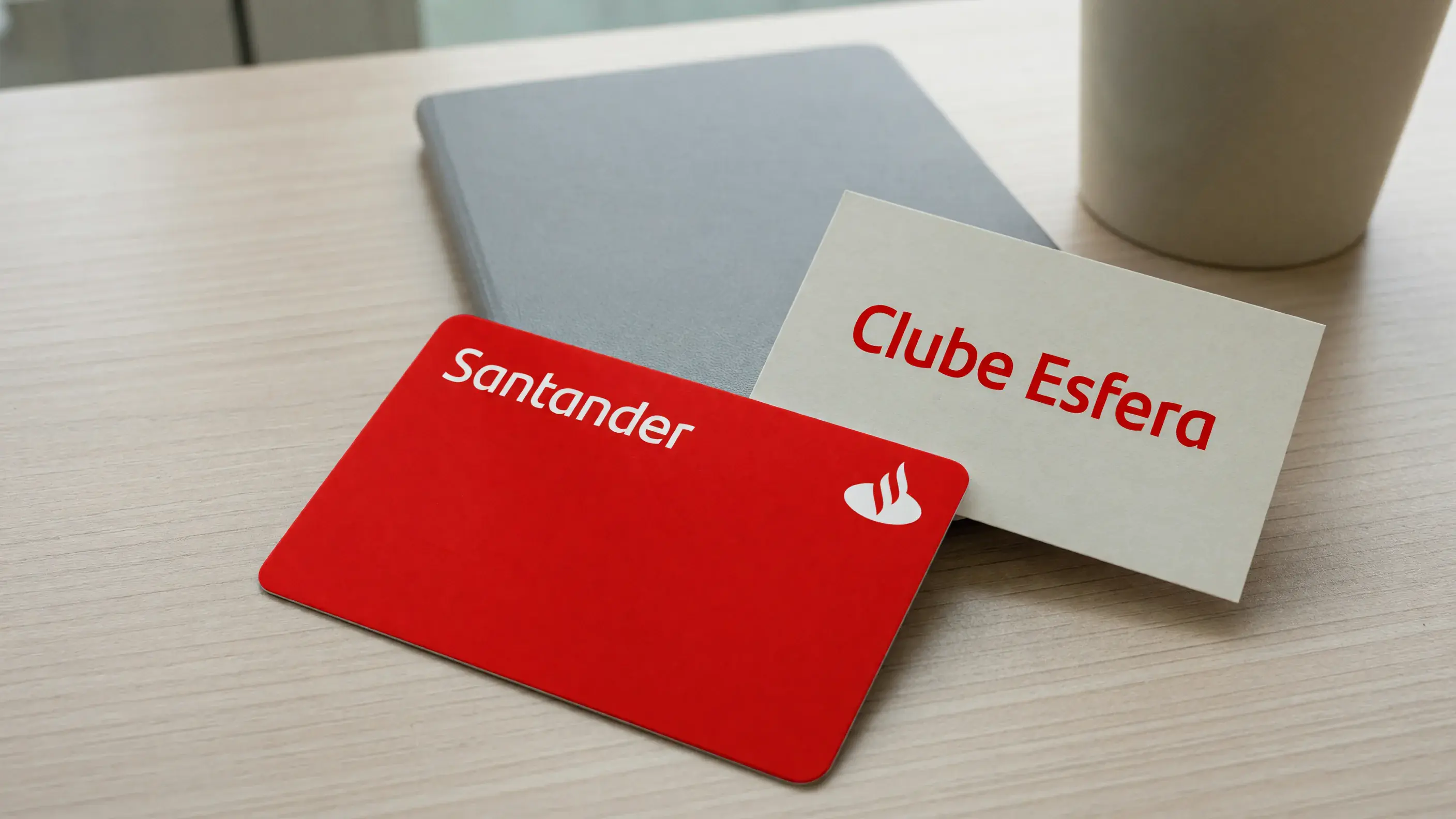 Cartão de crédito Santander vermelho em close diagonal sobre uma mesa de madeira clara de escritório, ao lado de uma placa de papel impressa com a marca-palavra Clube Esfera em vermelho.