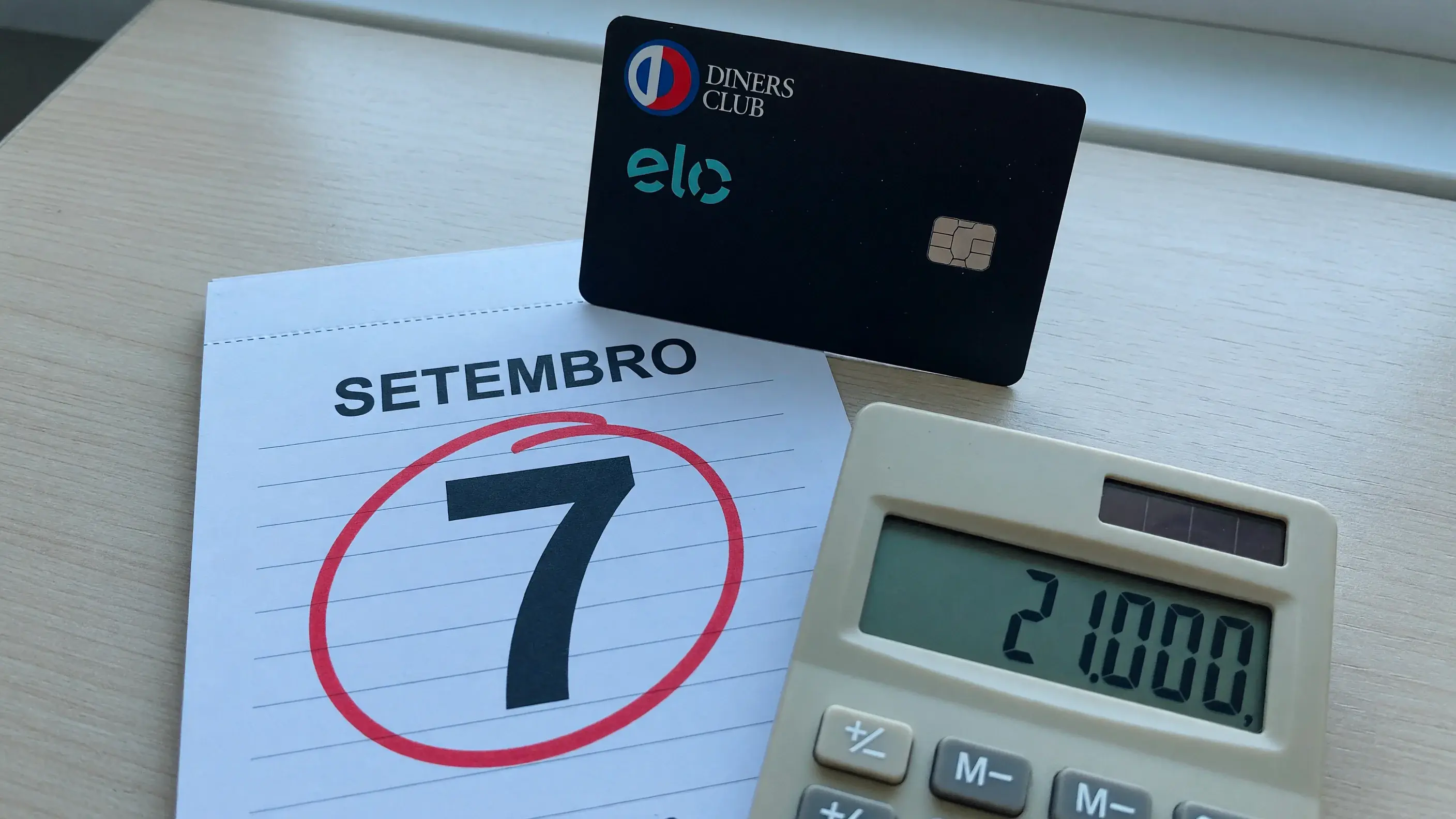 Cartão Elo Diners Club apoiado em pé sobre a folha de setembro de um calendário de mesa, com o dia 7 circulado em vermelho, e ao lado uma calculadora com 21.000 no visor — sem número de cartão legível.