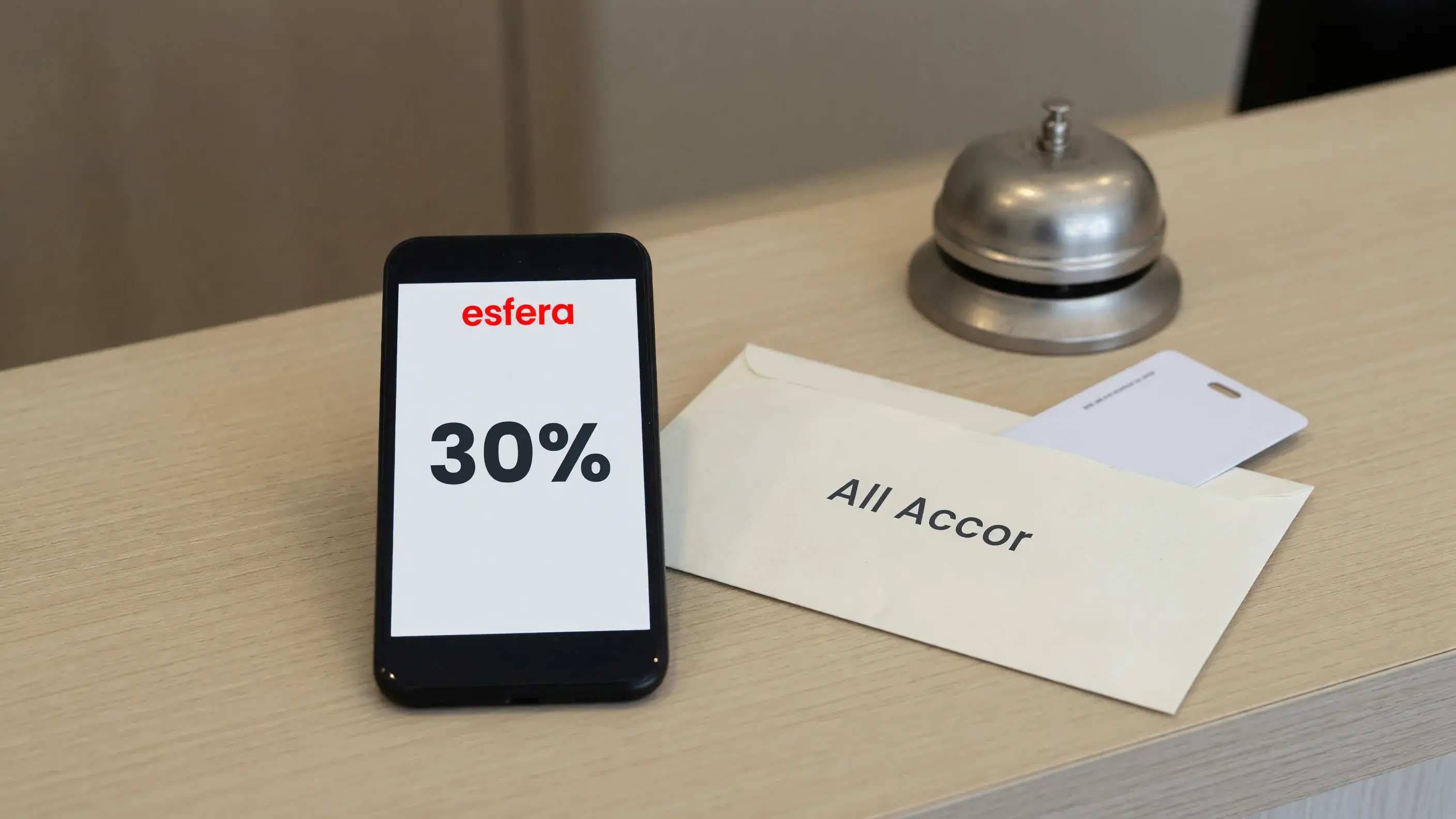 Celular apoiado no balcão de madeira de uma recepção de hotel, com a marca-palavra vermelha da Esfera e o número 30% na tela, ao lado de um cartão-chave de hotel num envelope de papel com o nome ALL Accor.