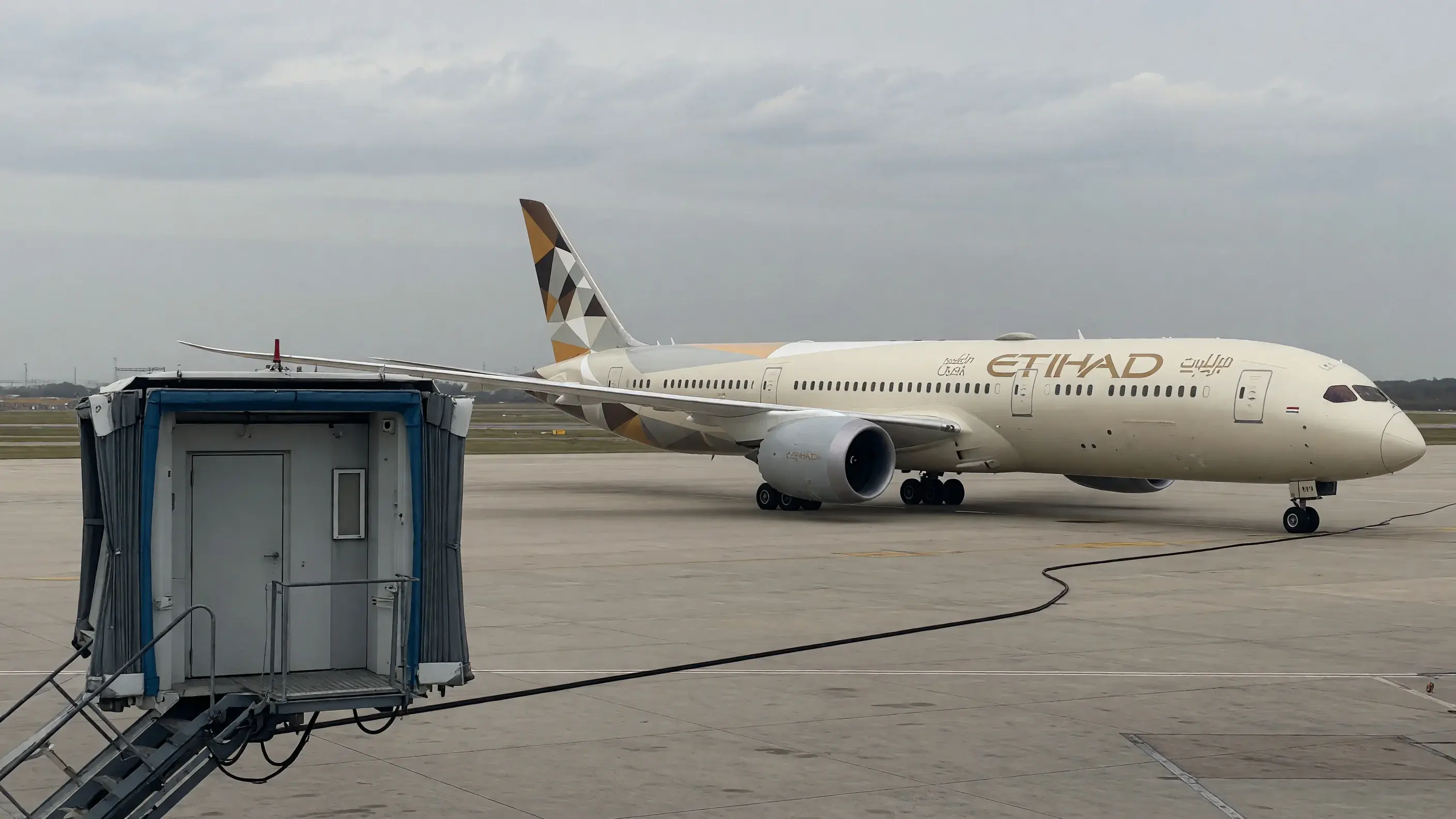 Avião da Etihad Airways, a companhia do programa Etihad Guest, parado no pátio sob céu encoberto, com a marca Etihad na fuselagem e na cauda, e a ponte de embarque recolhida e vazia em primeiro plano.