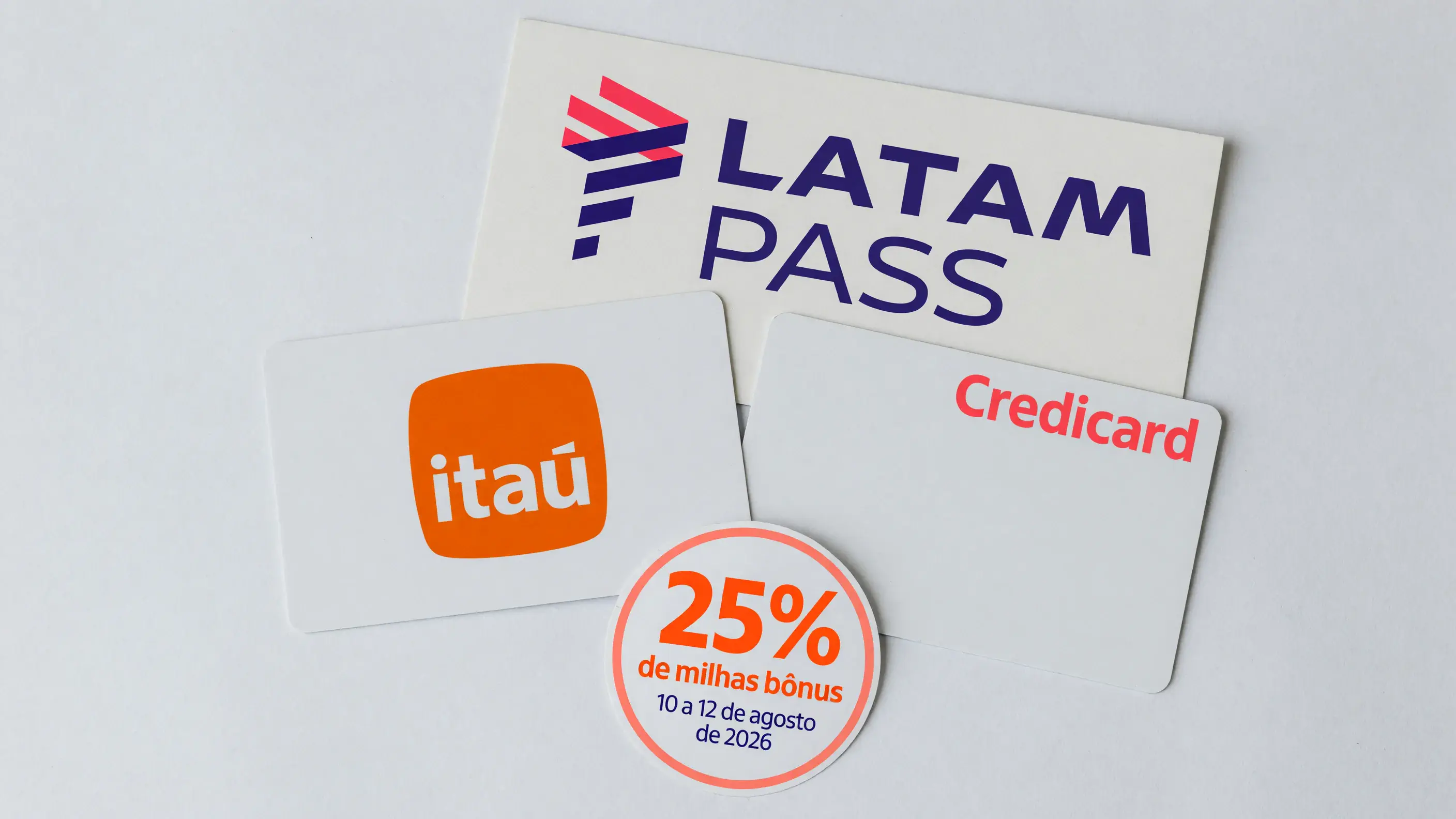 Dois cartões de crédito sobre uma bancada de papel, um com a marca Itaú e outro com a marca Credicard, ao lado de uma placa com a marca LATAM Pass e de um selo com 25% de milhas bônus e a janela de 10 a 12 de agosto de 2026.