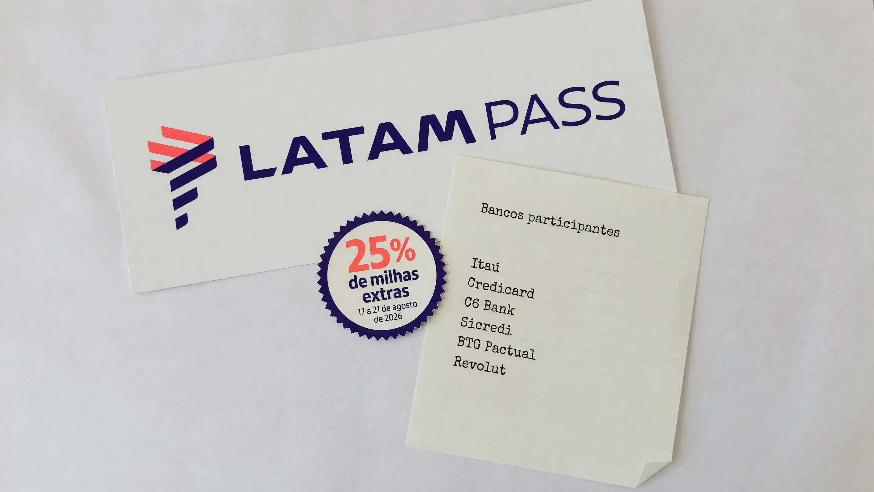 Placa impressa com a marca LATAM Pass sobre uma bancada de papel, ao lado de um selo com 25% de milhas extras e a janela de 17 a 21 de agosto de 2026, e de uma folha que lista em texto simples os bancos participantes da campanha.