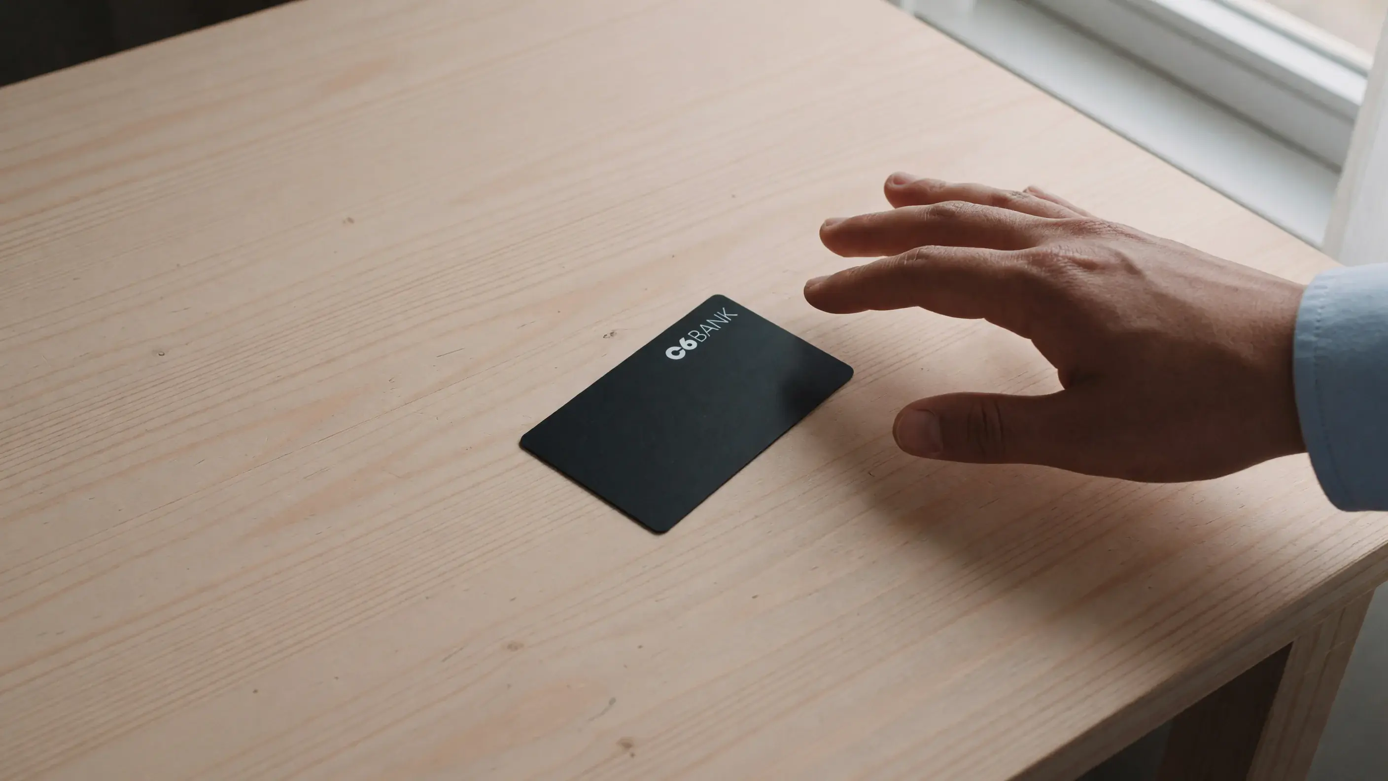 Cartão preto do C6 Bank sobre uma mesa de madeira clara, com a marca C6 Bank legível na face, e uma mão adulta se afastando do cartão sem pegá-lo.