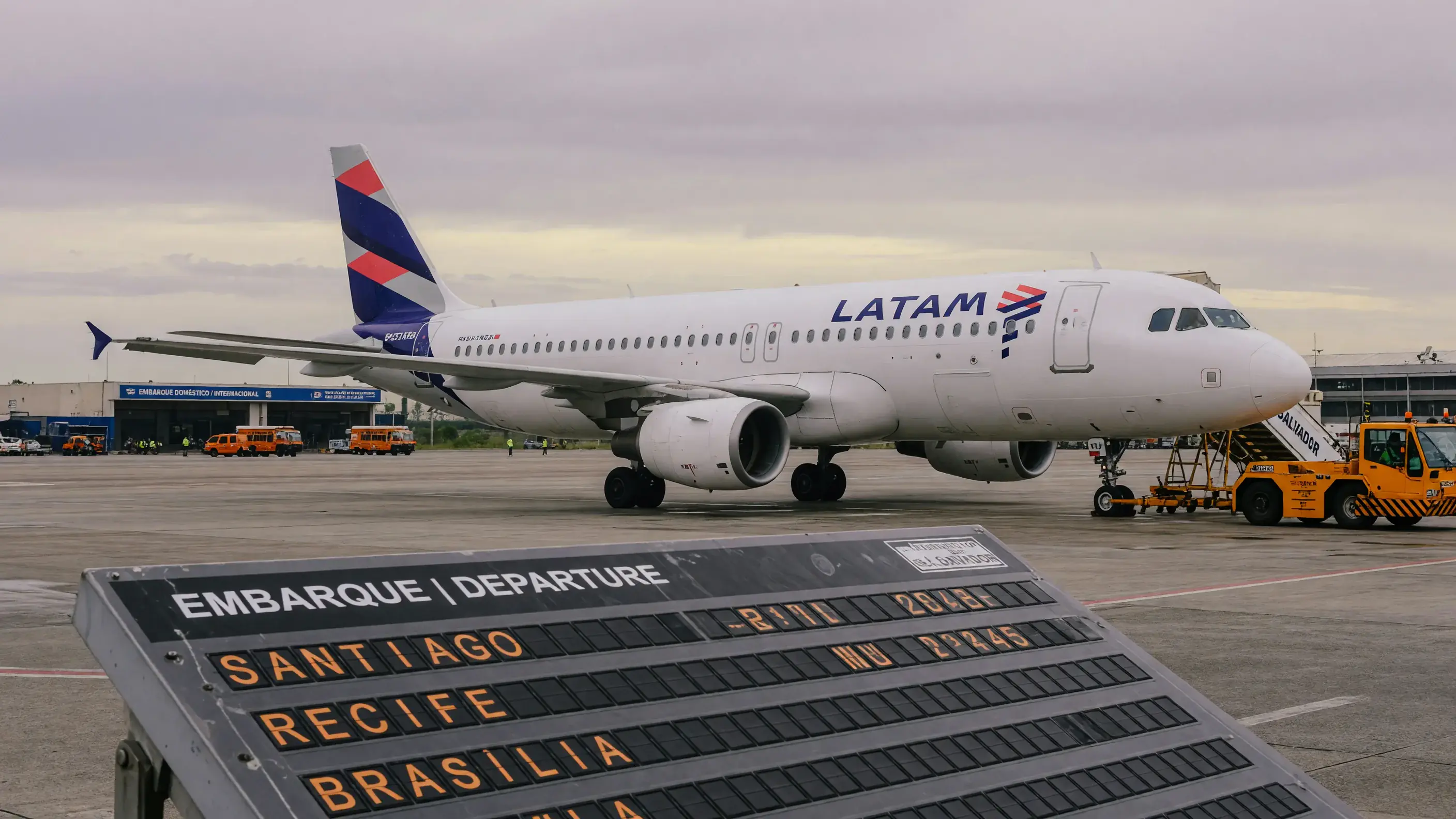 Avião com a pintura da LATAM parado no pátio do aeroporto de Salvador ao entardecer, com o painel de embarque indicando Santiago