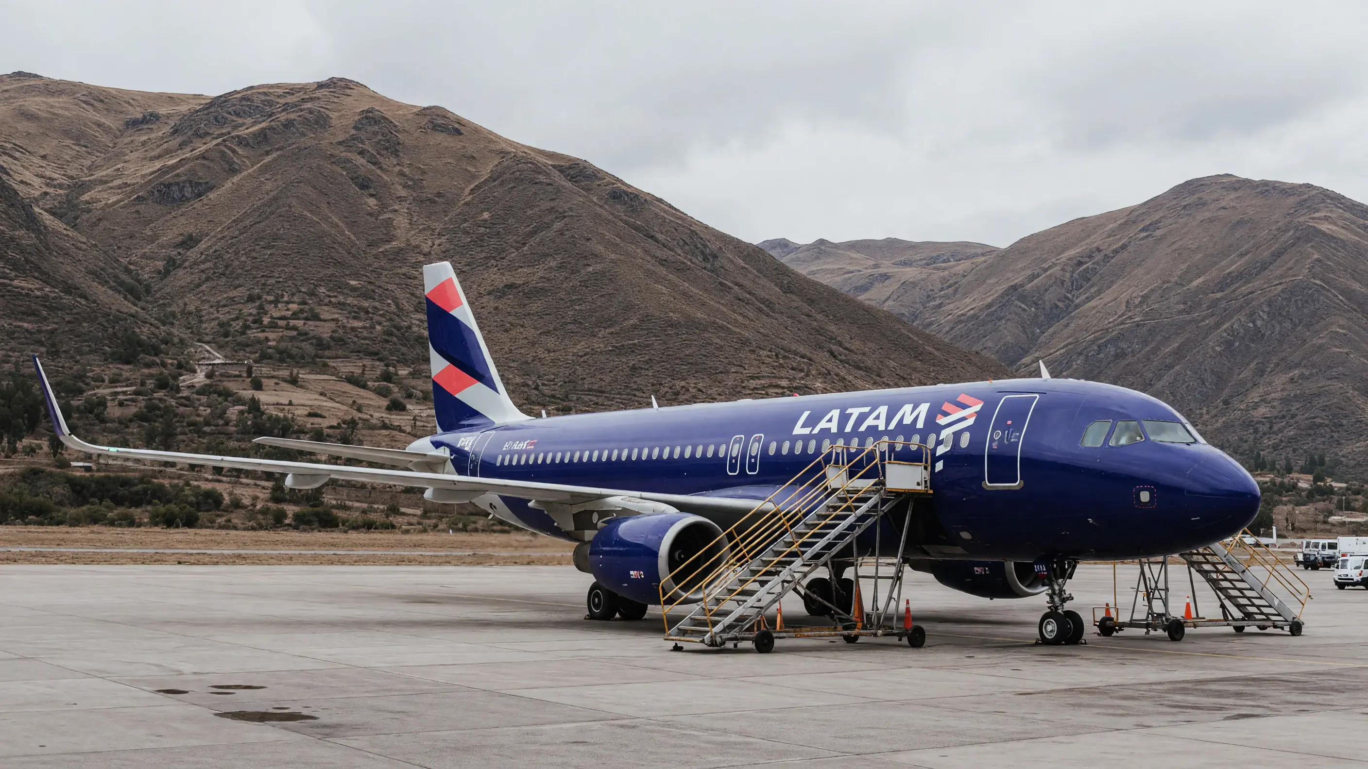 Aeronave da família Airbus A320 da LATAM parada no pátio de um aeroporto de montanha em Cusco, com a pintura índigo da companhia na fuselagem e o emblema em losango na deriva, a escada de embarque encostada na porta dianteira e as encostas secas dos Andes fechando o horizonte atrás do avião