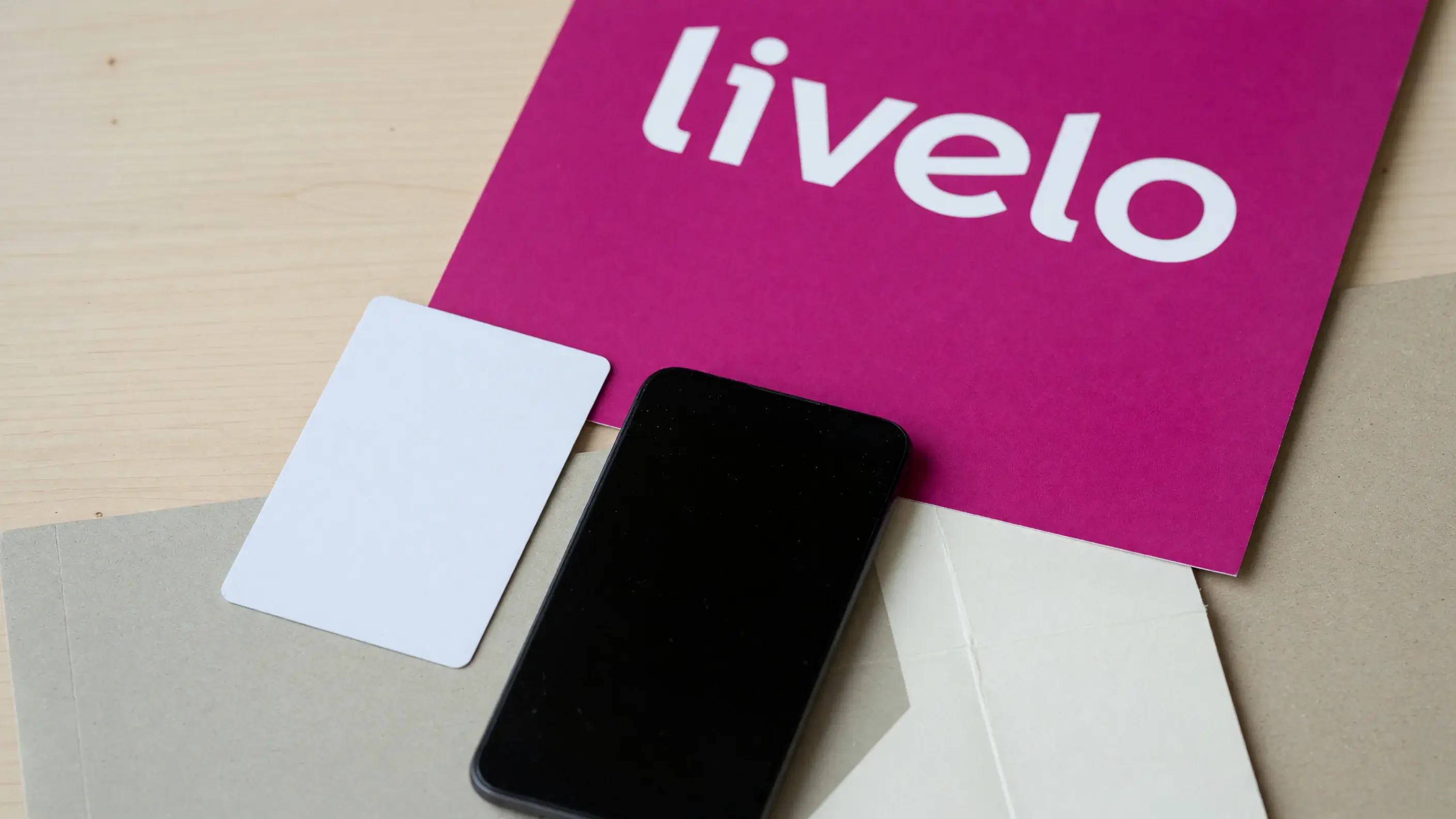 Bancada de madeira clara com um cartão de crédito sem marca e um celular de tela apagada, diante de um painel magenta com a marca-palavra livelo.