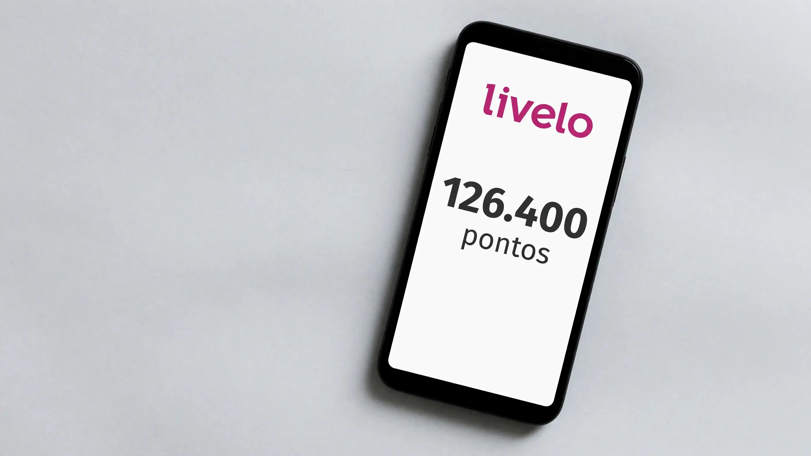 Celular sobre uma mesa de papel claro, com a marca da Livelo em magenta na tela e um campo de saldo de pontos em destaque.