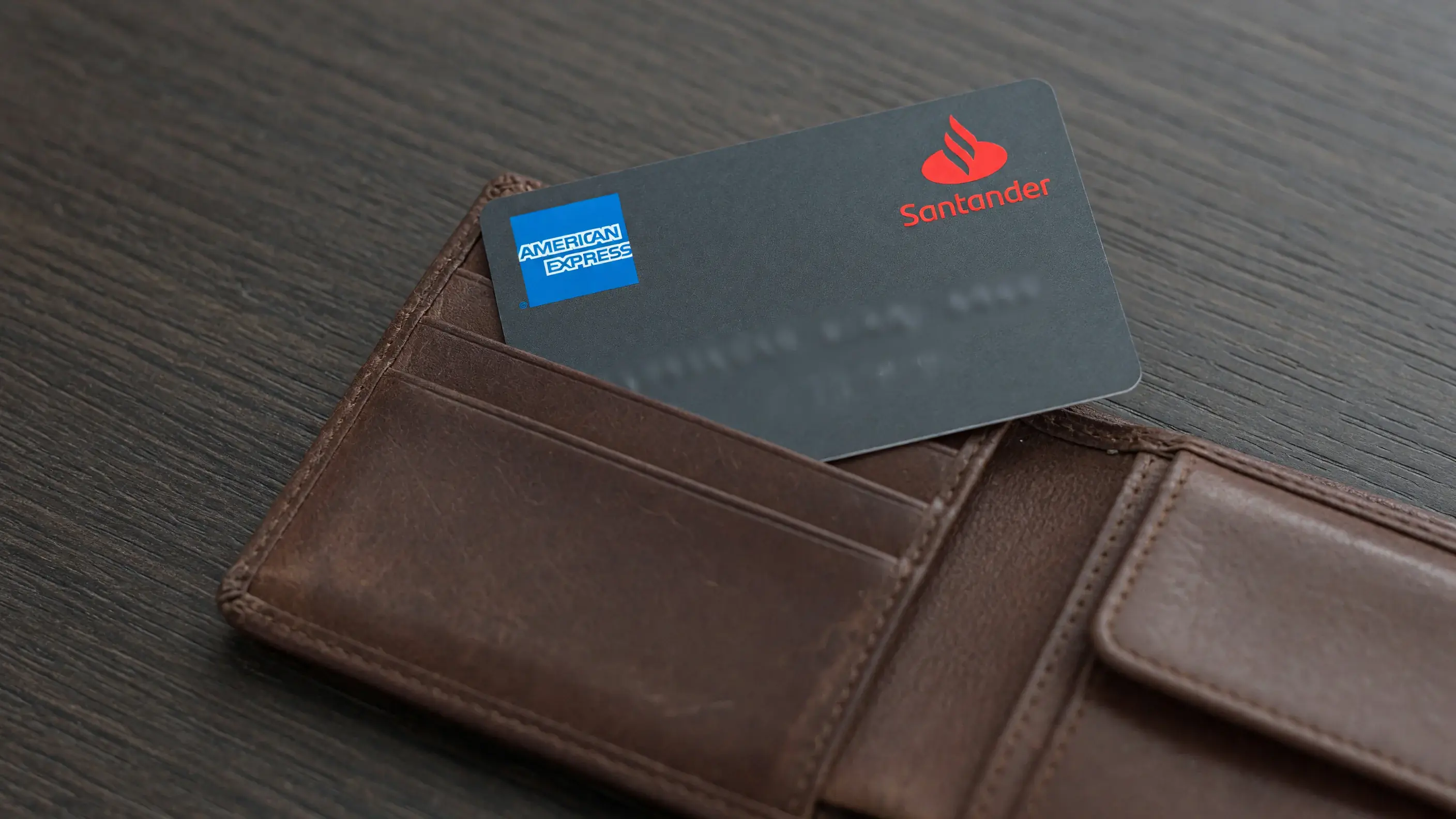 O cartão Amex do Santander, de face para cima e meio para fora de uma carteira de couro aberta sobre uma mesa de madeira escura, com a marca American Express e o logotipo do Santander nítidos na face.