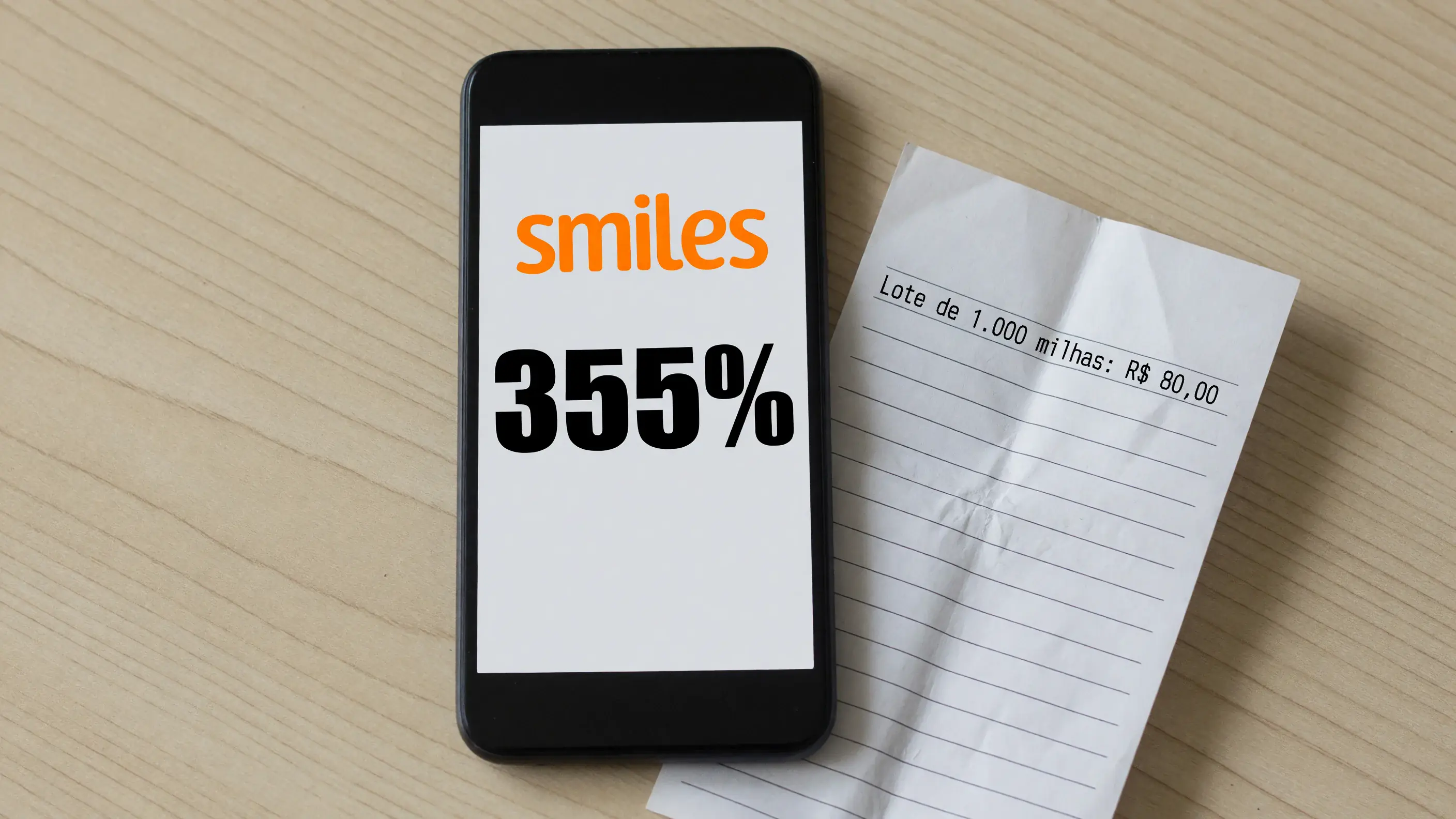Celular sobre uma mesa de madeira clara mostrando a marca-palavra laranja da Smiles e o número 355% na tela, ao lado de um recibo de papel com o preço do lote de mil milhas.