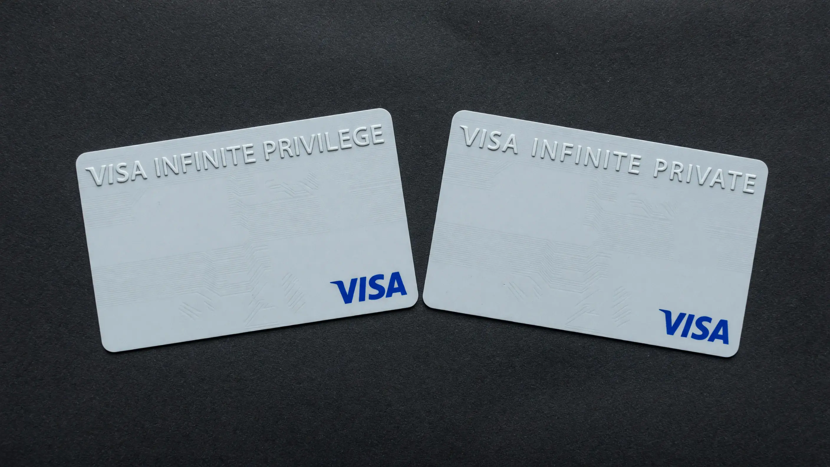 Dois cartões de crédito sem marca de banco, lado a lado sobre fundo escuro: à esquerda o plástico gravado Visa Infinite Privilege, à direita o mesmo desenho já gravado Visa Infinite Private, a troca de nome como único elemento diferente entre os dois.